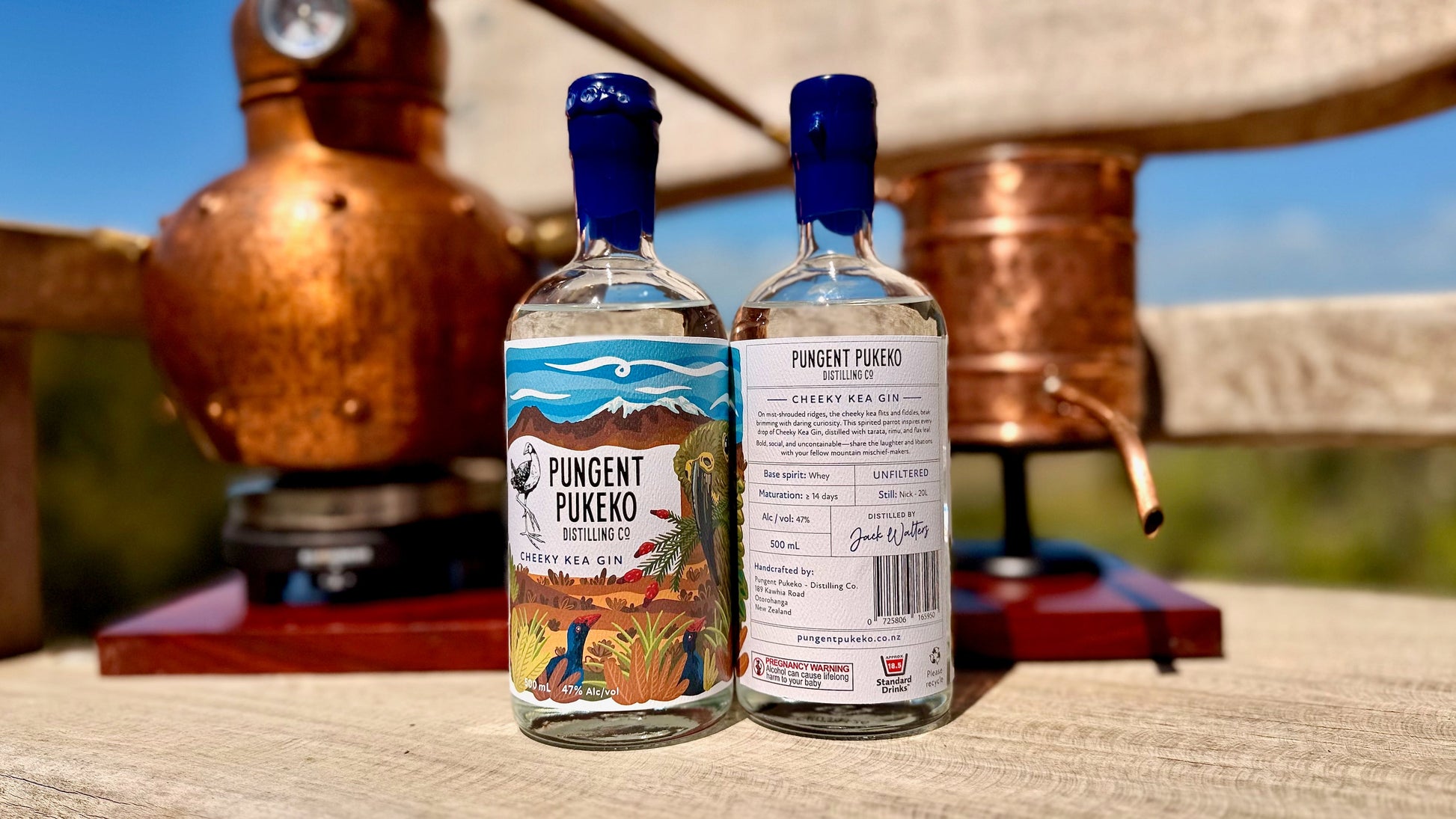 Two bottles of Cheeky Kea Gin with a scenic label, placed in front of copper stills.
